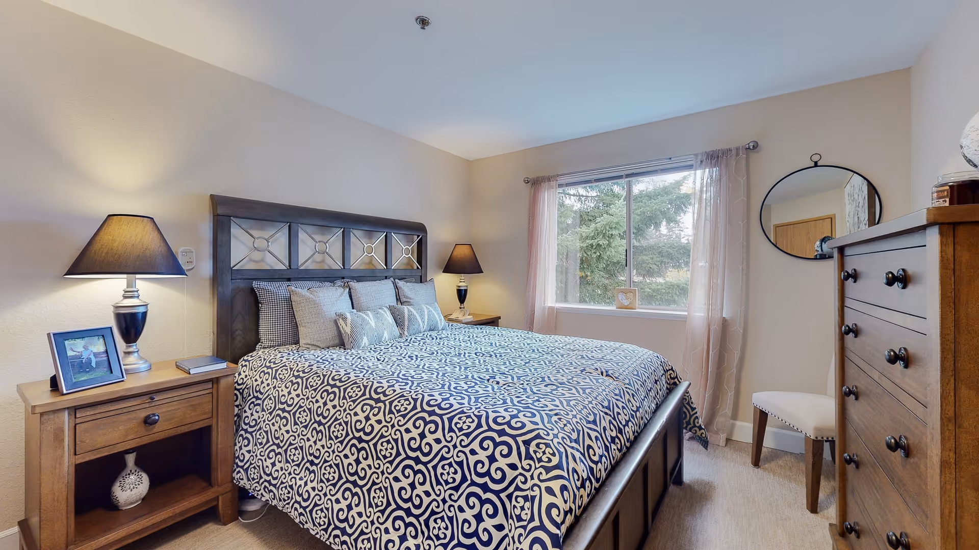 A cozy bedroom with a large bed covered in a blue and white patterned comforter and multiple pillows. There are two wooden nightstands on either side of the bed, each with a lamp. A window with sheer curtains lets in natural light, and a round mirror hangs on the wall above a wooden dresser. A small cushioned chair is placed next to the dresser.