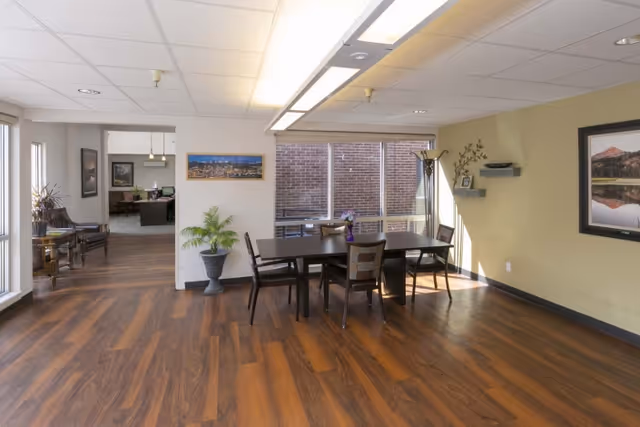 A well-lit interior room with wood flooring featuring a rectangular dining table with six chairs. There is a potted plant near the wall, framed pictures on the walls, and large windows letting in natural light. In the background, there is an office area with desks and chairs.