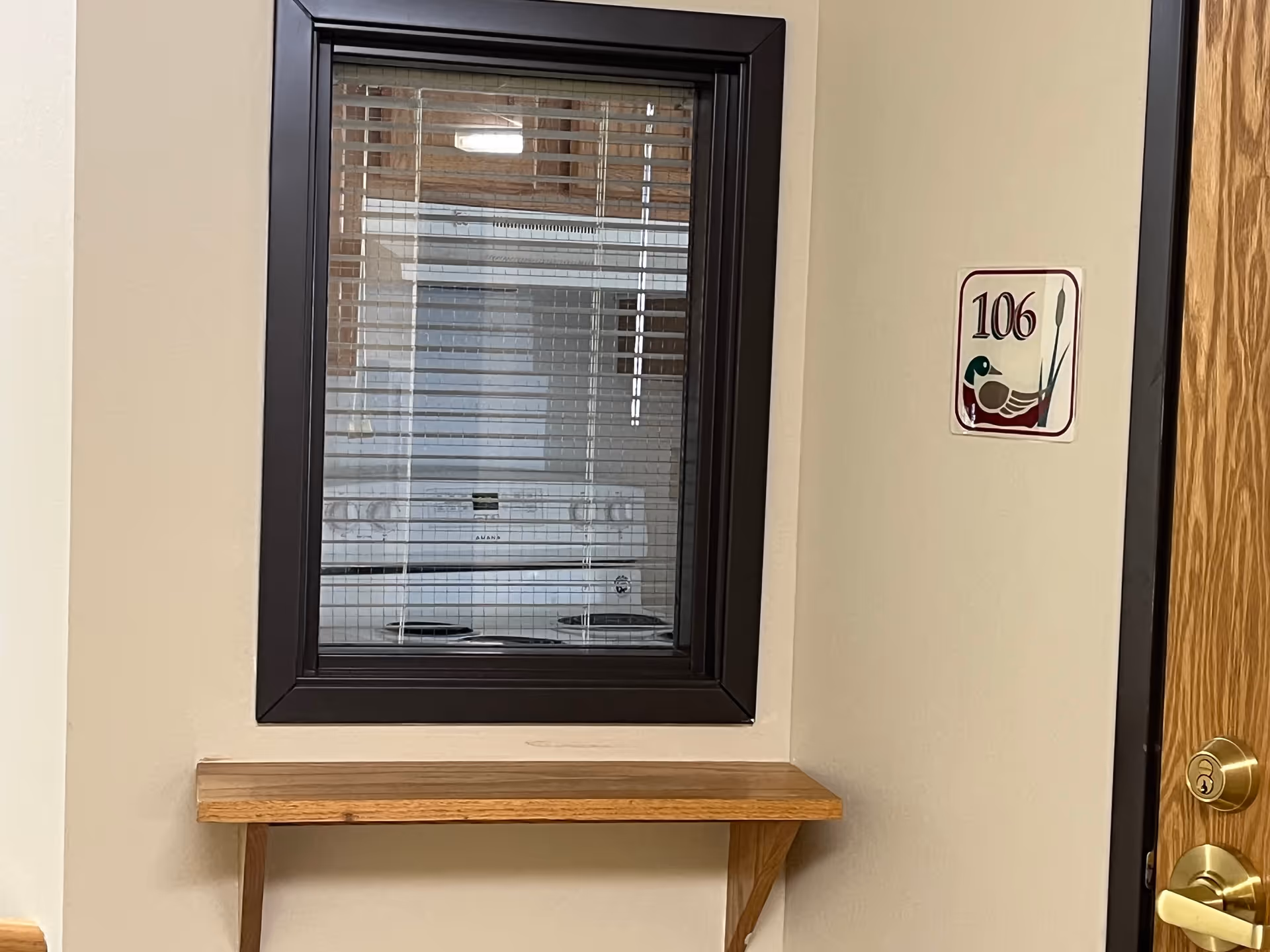 Interior view of a room with a window featuring horizontal blinds, a small wooden shelf mounted on the wall below the window, and a door with a gold-colored lock and handle. A room number sign '106' with a duck illustration is mounted on the wall next to the door.