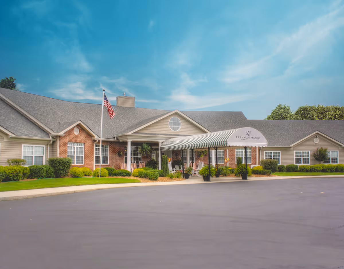 Exterior front view of Franklin Manor, a single-story building with a gray roof, beige siding, and brick accents. The entrance features a covered awning with the Franklin Manor logo and name, surrounded by neatly trimmed bushes and an American flag on a flagpole. The sky is clear with some clouds.