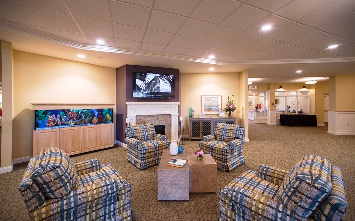A cozy senior living lounge with patterned armchairs arranged around a coffee table, a wall-mounted TV above a fireplace, and an aquarium.