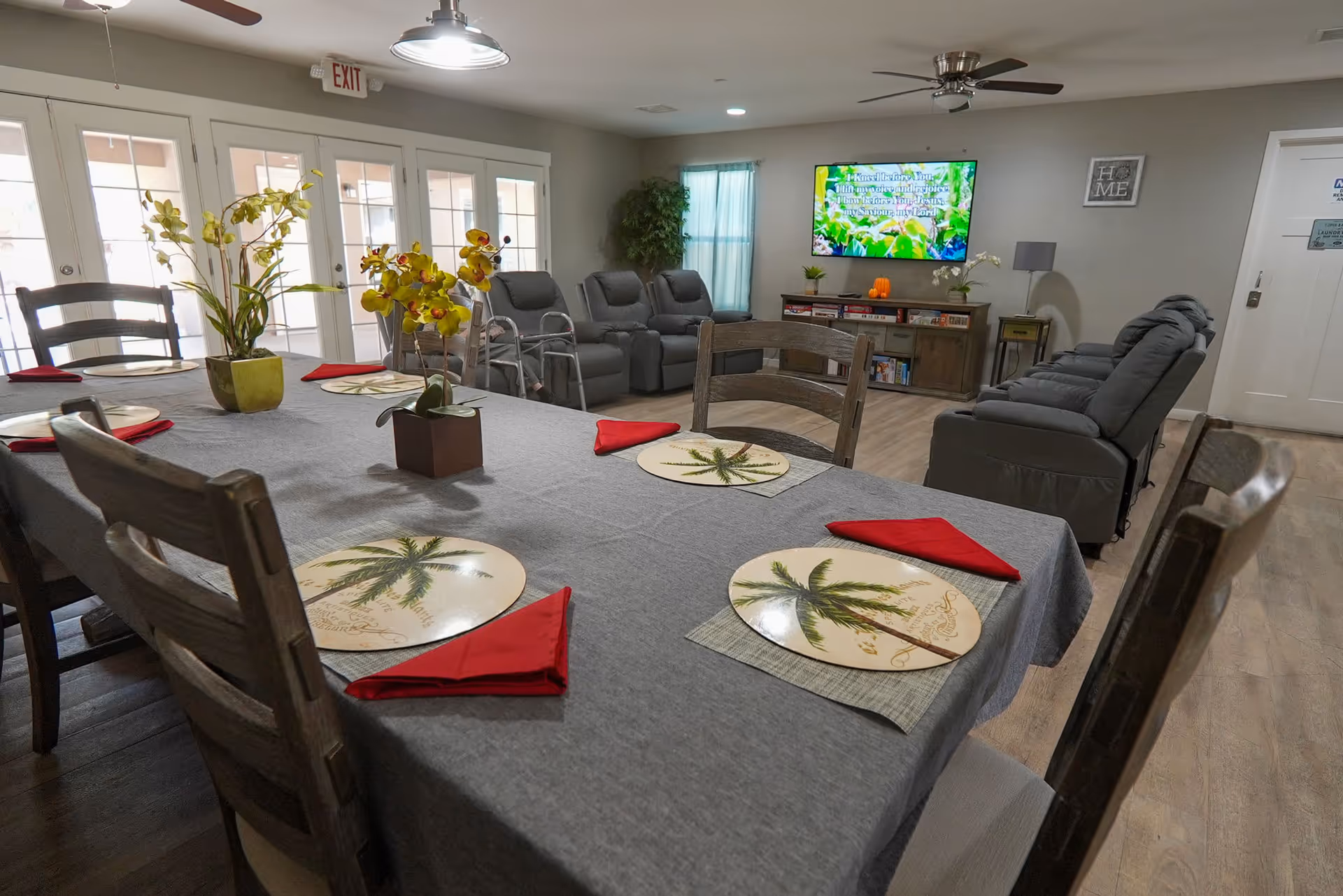 A communal living and dining area with a long table covered by a gray tablecloth, set with palm tree-themed placemats and red napkins. There are two potted orchid plants on the table. In the background, there are several gray recliner chairs arranged facing a wall-mounted TV displaying colorful text and images. The room has wooden flooring, ceiling fans, and large glass doors letting in natural light.