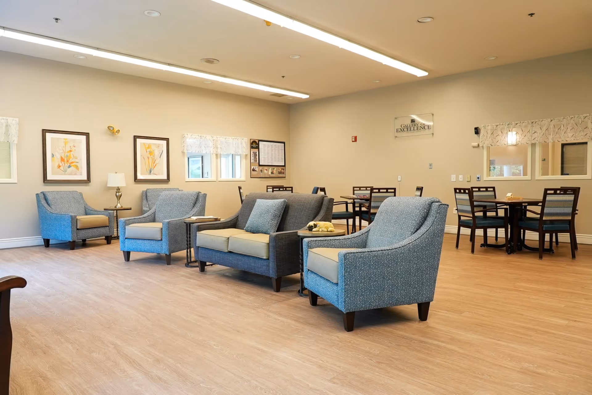 A spacious and well-lit common area with light wood flooring, featuring several upholstered armchairs and a loveseat arranged around small side tables. In the background, there are dining tables with chairs, framed floral artwork on the walls, windows with valances, and a sign that reads 'Gallery of Excellence'.