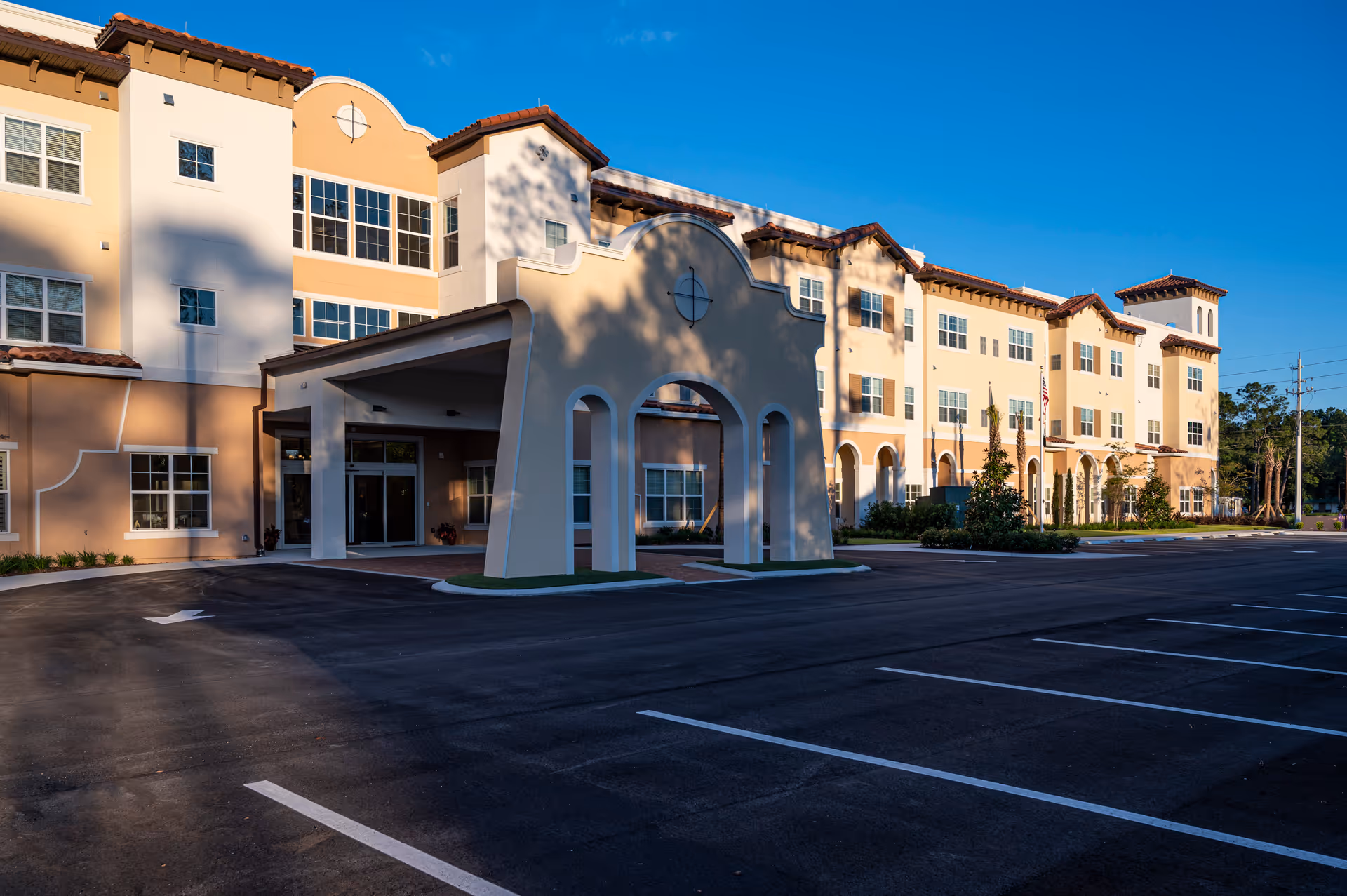 Exterior view of a multi-story senior living facility building with a covered entrance and an empty parking lot under a clear blue sky.