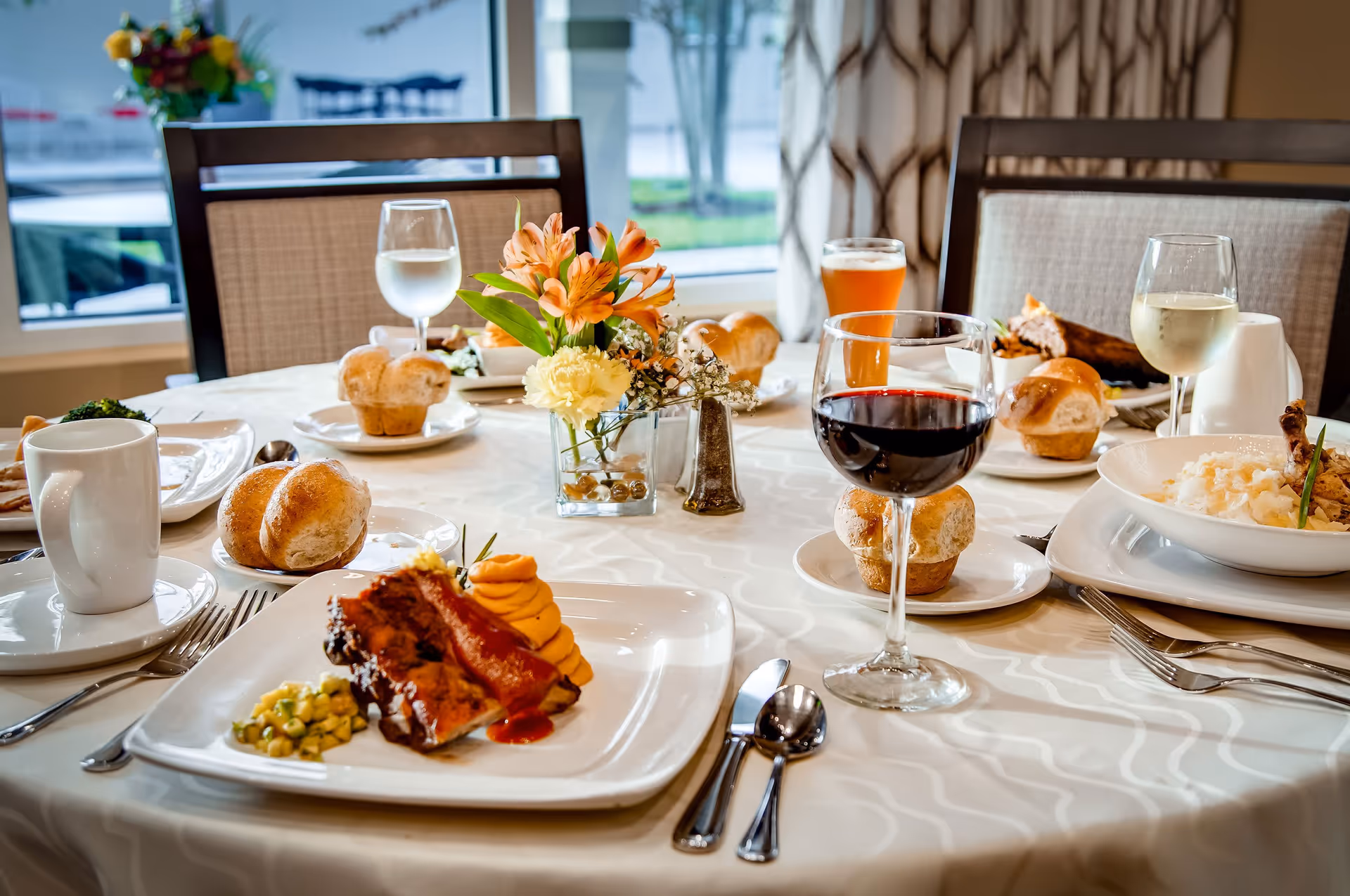 A dining table set with plates of food including ribs with sauce, mashed sweet potatoes, rolls, and a bowl of rice with chicken. There are glasses of red wine, white wine, water, and a beer on the table. A small flower arrangement with orange and yellow flowers is in the center of the table. The background shows a window with a view outside and patterned curtains.