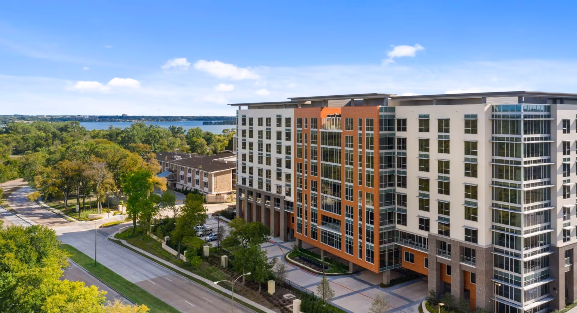 Aerial view of a modern multi-story senior living facility building with large windows, surrounded by trees and greenery, with a road running alongside it and a body of water visible in the background under a clear blue sky.