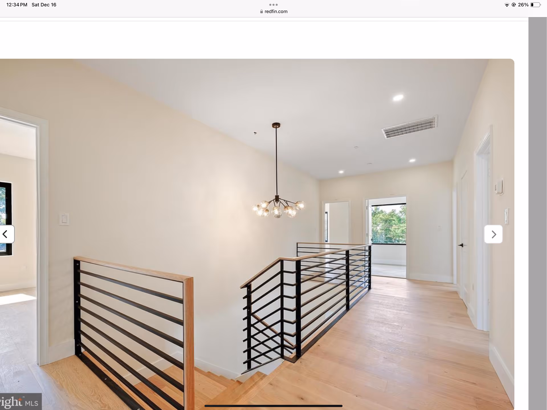 Interior view of a hallway with light wood flooring and beige walls. A modern black metal and wood railing lines the staircase. A contemporary chandelier with multiple exposed bulbs hangs from the ceiling. Doors and windows are visible at the end of the hallway, allowing natural light to enter.