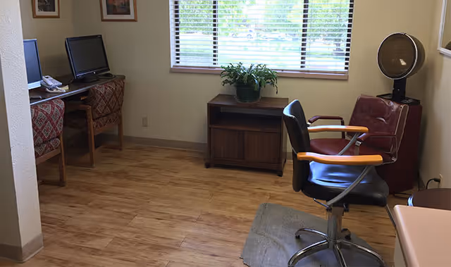 Interior room with salon chairs (one under a hair dryer), a computer workstation with two monitors, a small cabinet, and a window with blinds.