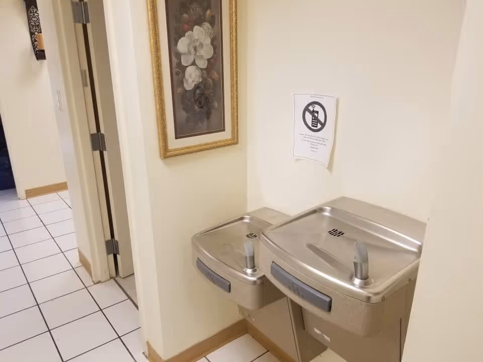 Two stainless steel drinking fountains mounted on a beige wall in a tiled hallway. Above the fountains, there is a sign with a no cell phone symbol. A framed floral picture hangs on the adjacent wall near an open door.