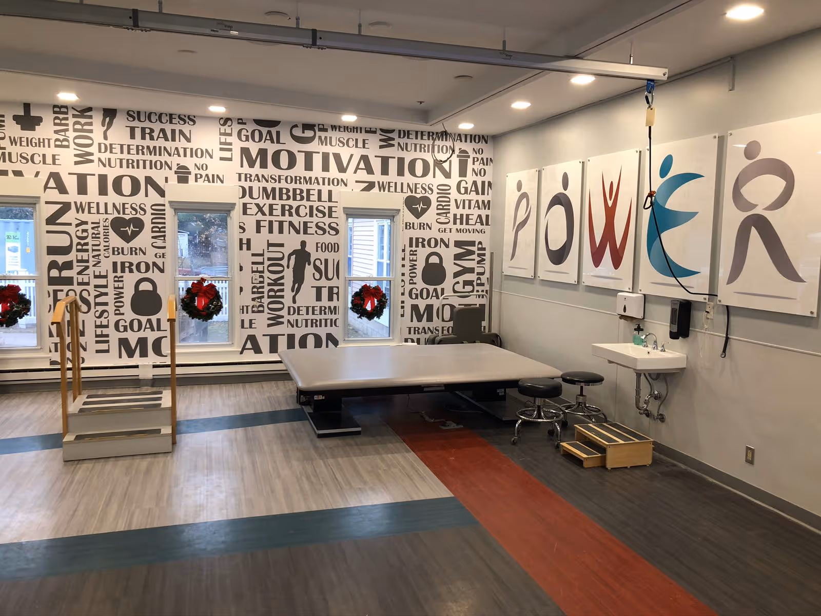 A rehabilitation room with a large padded therapy table, two stools, a small sink, and a wooden step platform. The walls are decorated with motivational words related to fitness and wellness, and five large panels spelling out the word 'POWER' with stylized human figures. Three windows have holiday wreaths hanging on them.