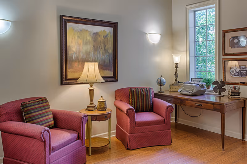 A cozy sitting area with two red upholstered armchairs featuring striped cushions, a small round wooden side table with a lamp and decorative items, and a wooden desk with a vintage typewriter, globe, lamp, and fan. A large window lets in natural light, and framed artwork hangs on the walls.