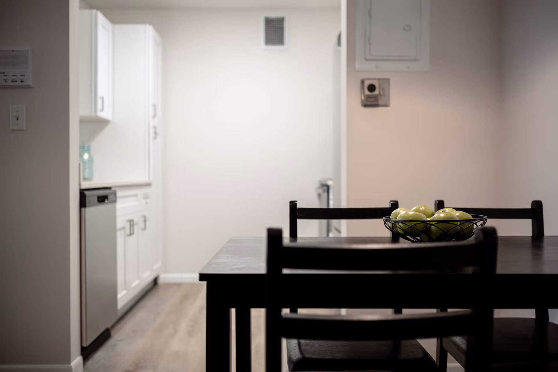 View of a dining area with a dark wooden table and chairs. A bowl of green apples is placed on the table. In the background, there is a kitchen with white cabinets and a dishwasher.