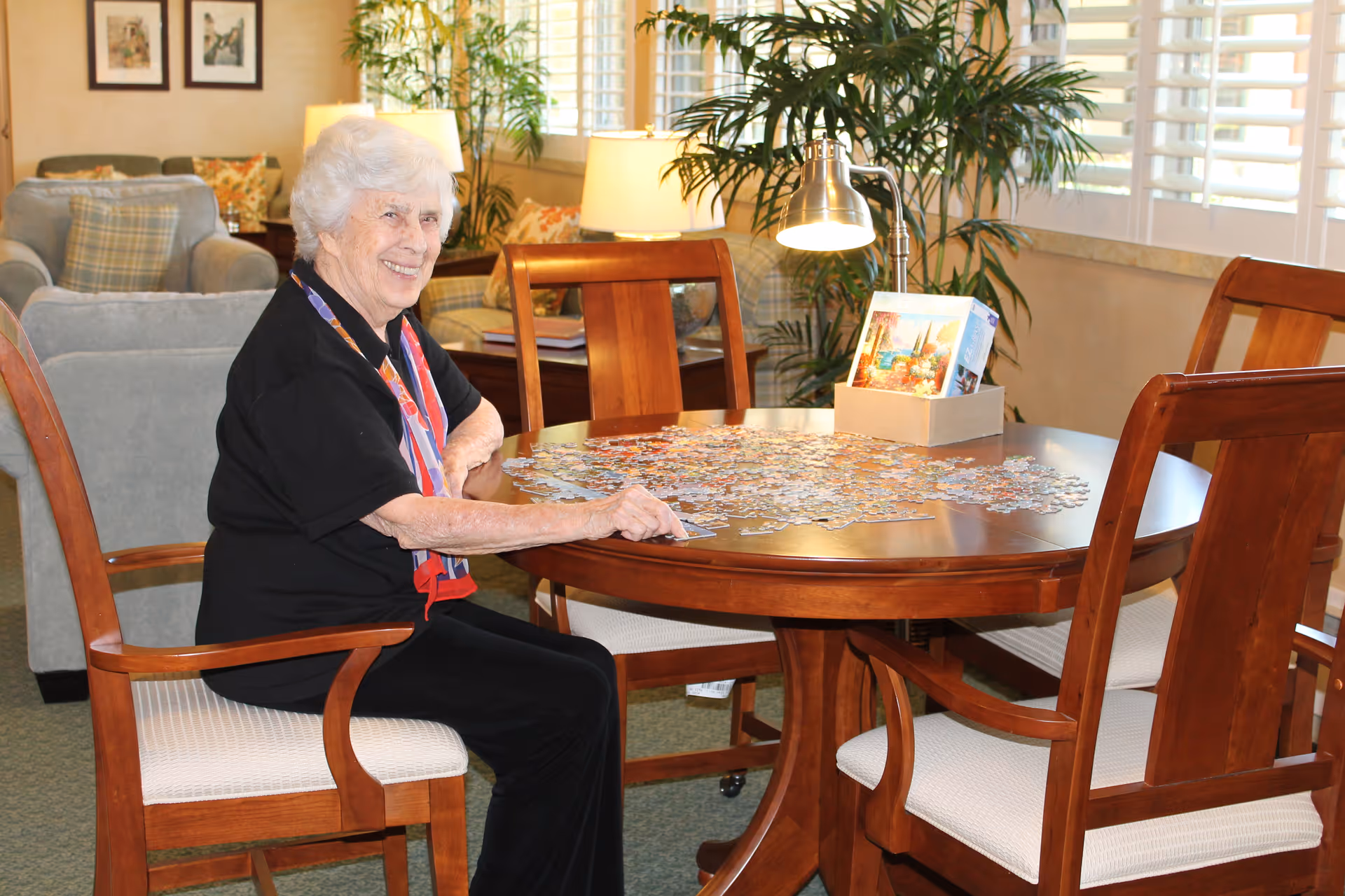 An elderly woman with white hair is sitting at a round wooden table working on a jigsaw puzzle in a cozy living room area. The room has comfortable chairs, a lamp, framed pictures on the wall, and large windows with white shutters letting in natural light. There are plants and decorative pillows adding warmth to the space.
