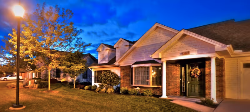 Exterior view of a senior living facility at dusk with well-lit houses, a street lamp, and a tree with autumn leaves. The houses have brick and siding exteriors with front porches and windows.