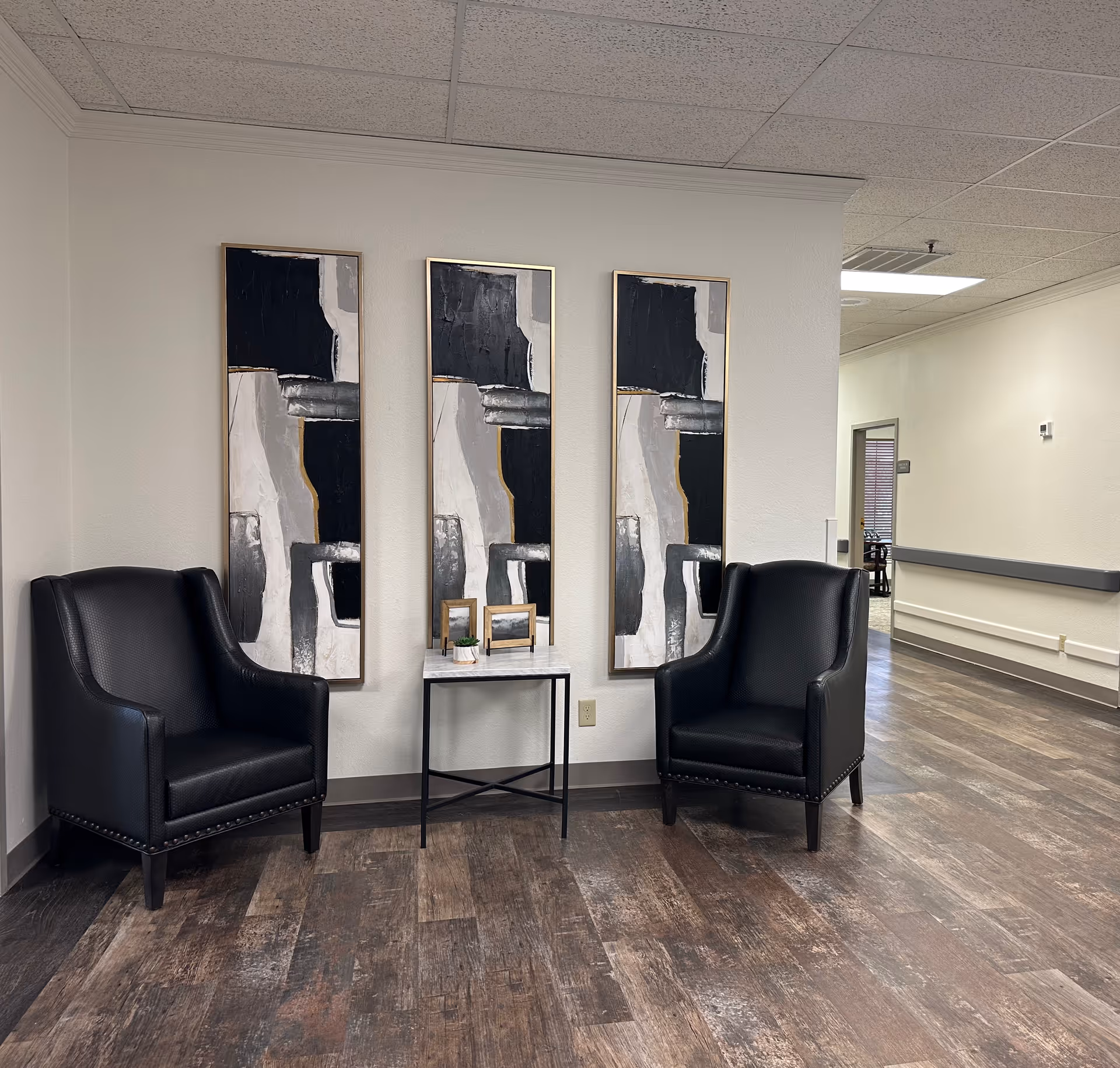 A seating area in a hallway with two black leather armchairs on either side of a small table. The table holds two small framed pictures and a small plant. Behind the table are three vertical abstract paintings with black, white, and gray tones. The floor is wood-patterned, and the walls are light-colored with a drop ceiling above.