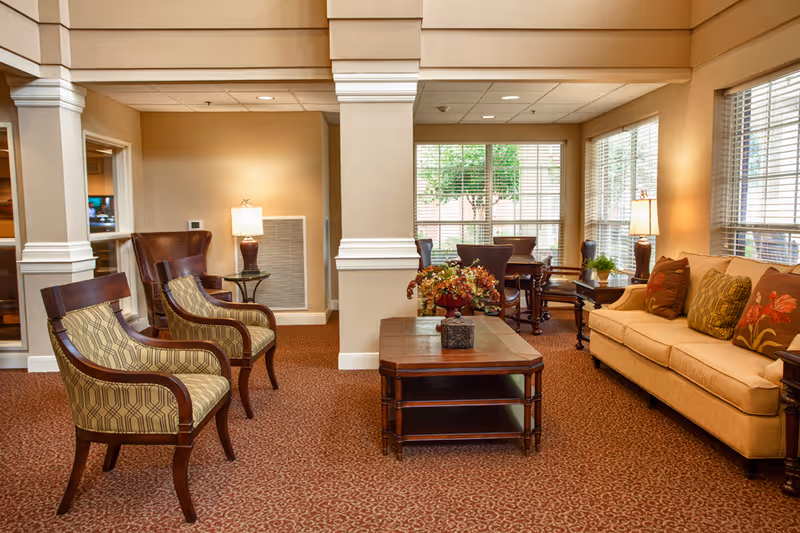 A cozy senior living facility common area with a beige sofa adorned with decorative pillows, two patterned armchairs, a wooden coffee table with a floral centerpiece, and large windows letting in natural light. The room has warm beige walls, carpeted floors, and table lamps providing additional lighting.