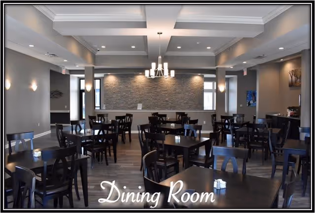A spacious dining room with multiple dark wooden tables and chairs arranged neatly. The room features a modern chandelier hanging from a coffered ceiling, soft wall lighting, and a textured stone accent wall at the far end. Large windows allow natural light to enter, and the floor is covered with wood-style flooring.