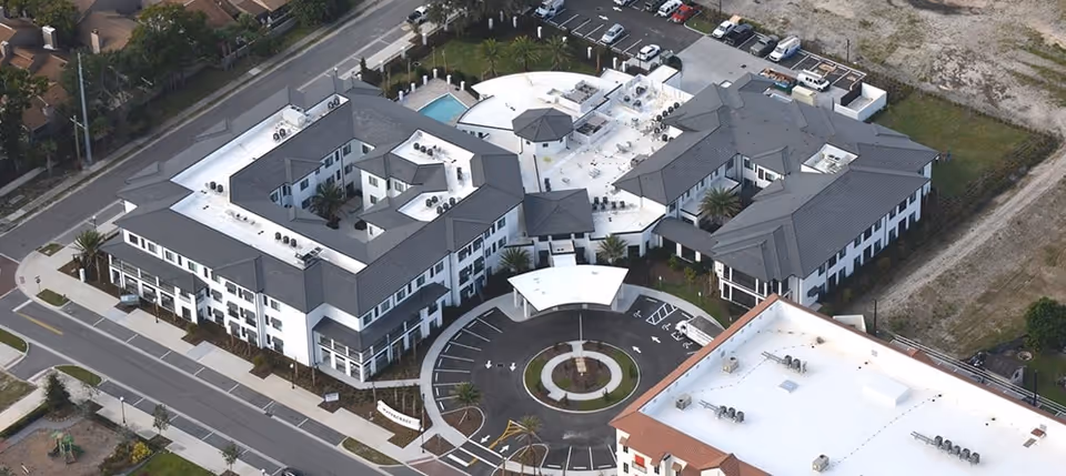 Aerial view of Watercrest Winter Park senior living facility showing multiple connected buildings with gray roofs, a circular driveway with a covered entrance, a swimming pool, and surrounding parking areas and streets.