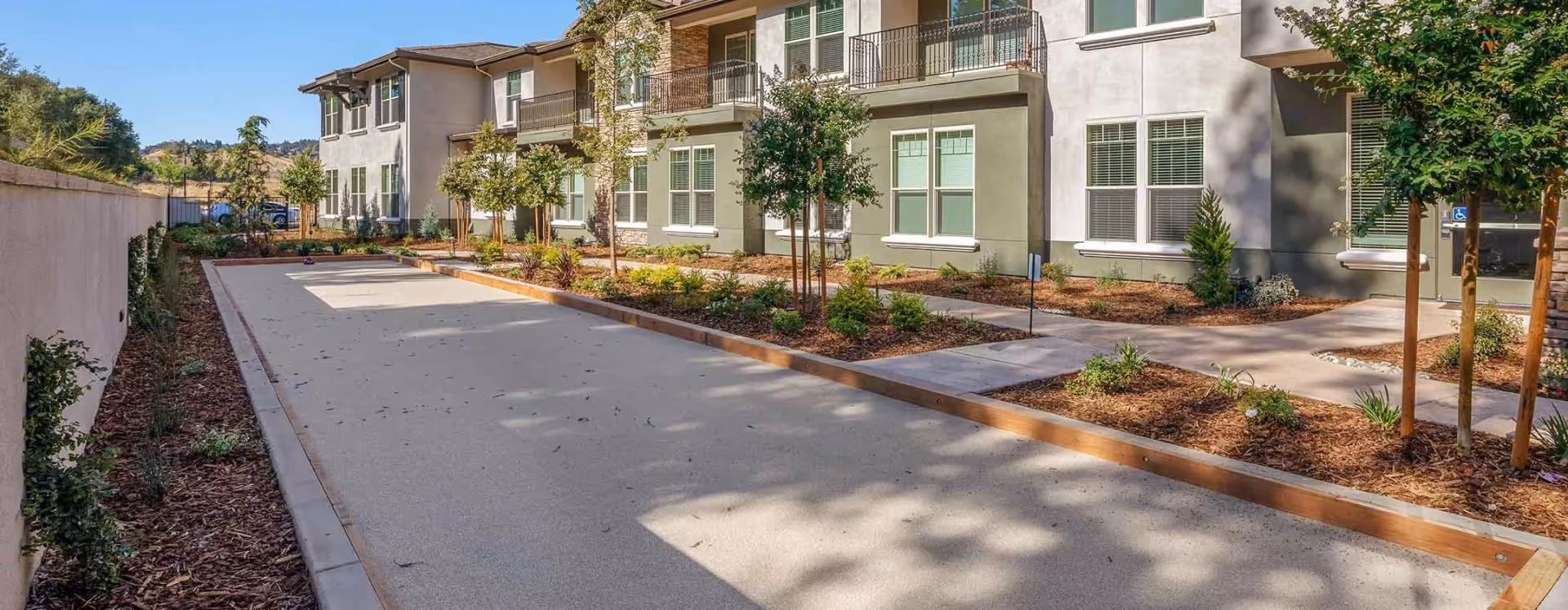 Outdoor bocce ball court with wooden borders surrounded by landscaped garden beds and young trees, adjacent to a two-story residential building with balconies and windows.