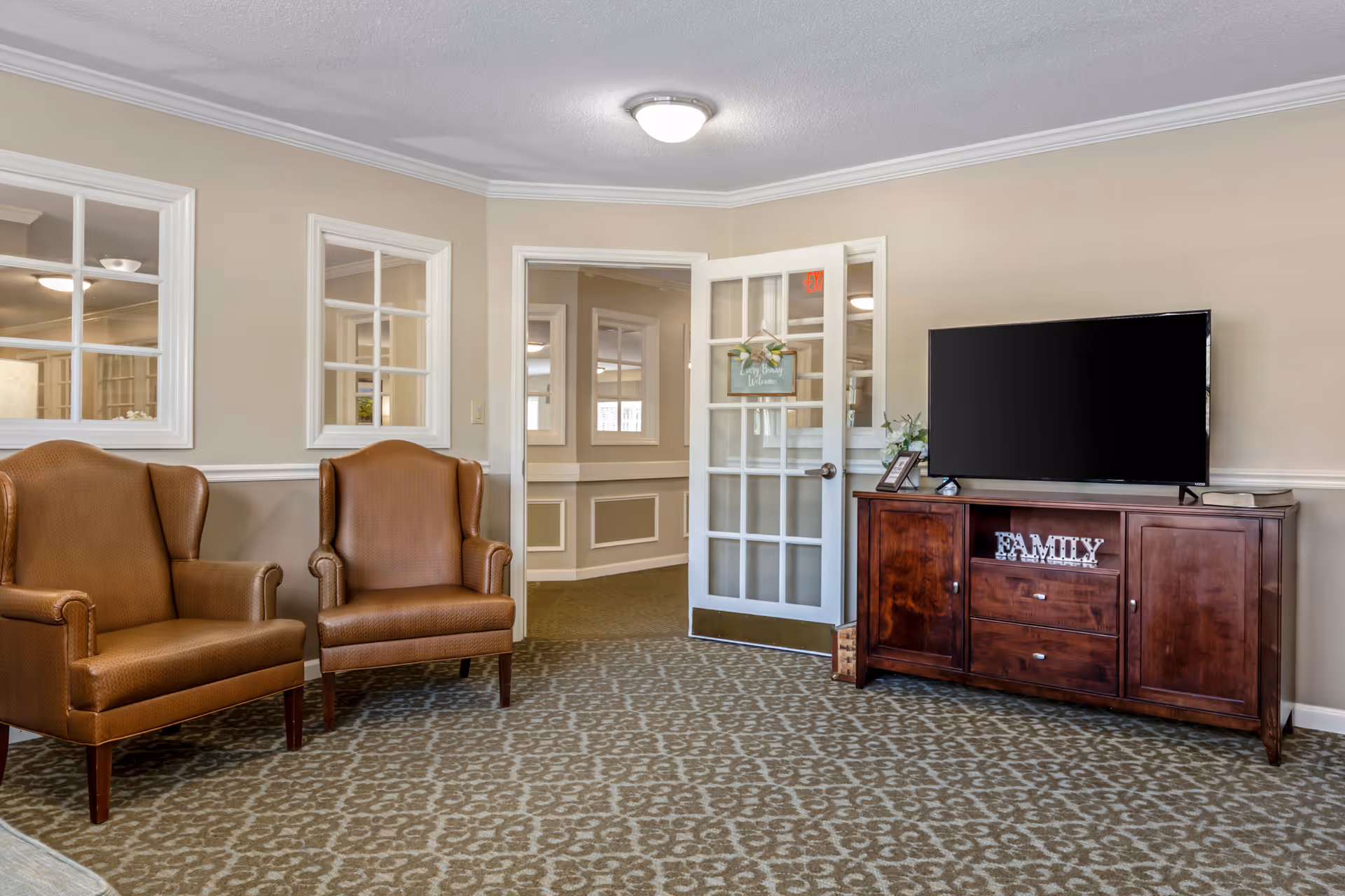 A cozy living room area with two brown leather armchairs on the left and a wooden TV stand on the right holding a flat-screen television and decorative items including a sign that says 'FAMILY'. The room has beige walls, patterned carpet, and a glass-paneled door that is open, showing a hallway with similar decor.