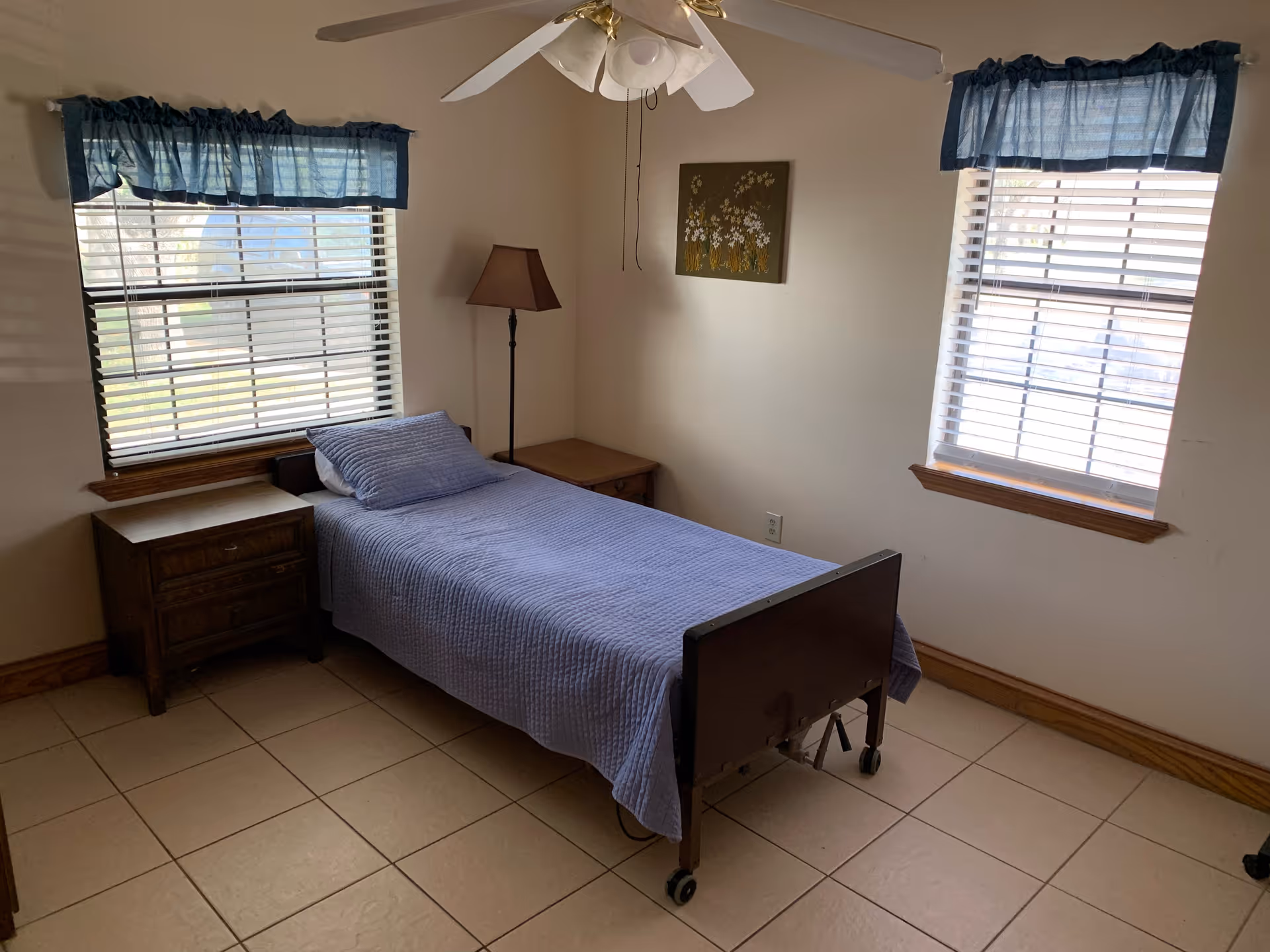 A simple bedroom with a single bed covered in a blue quilt and matching pillow. The room has two windows with blue valances, a ceiling fan with lights, a floor lamp, two wooden nightstands, and a small floral painting on the wall.