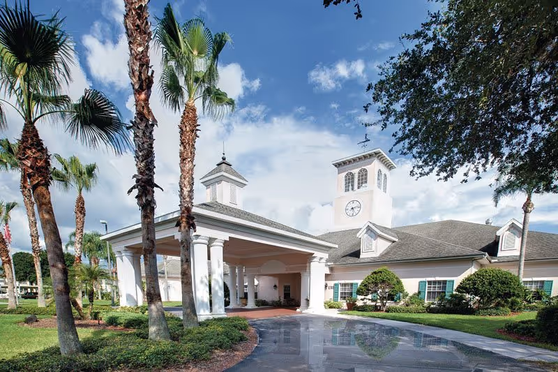 Front exterior of a senior living building with a porte-cochere, clock tower, and palm trees.