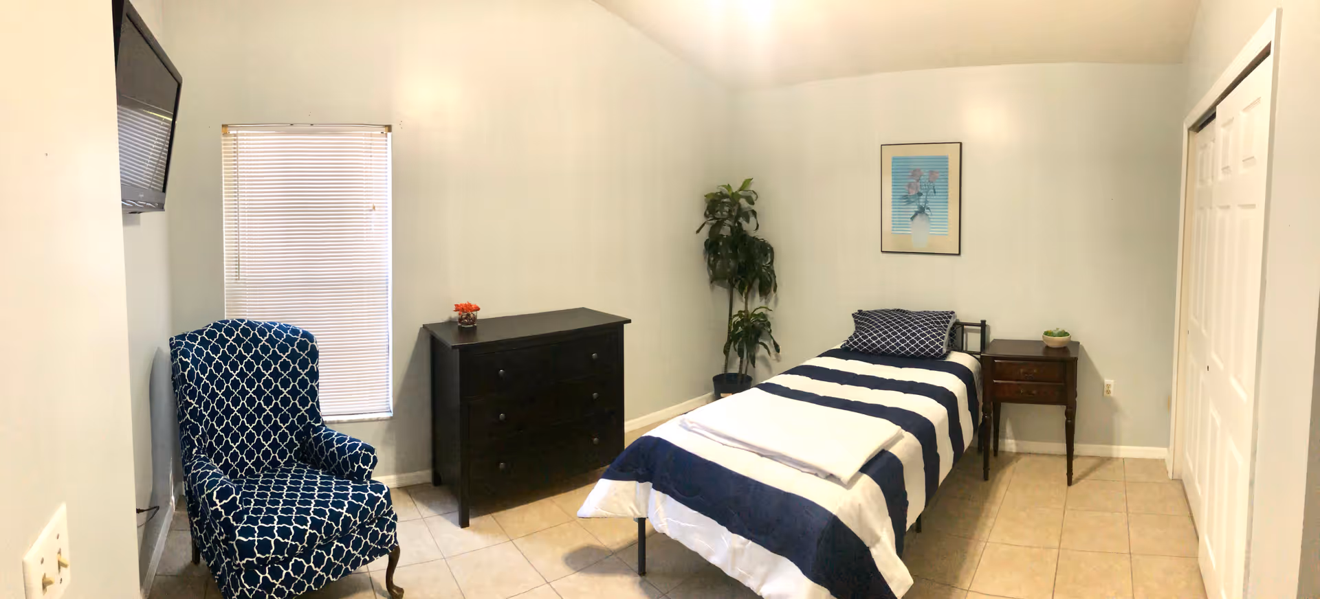 Private bedroom with a single bed dressed in navy-and-white striped bedding, a nightstand and dresser, upholstered chair, wall-mounted TV and a potted plant.