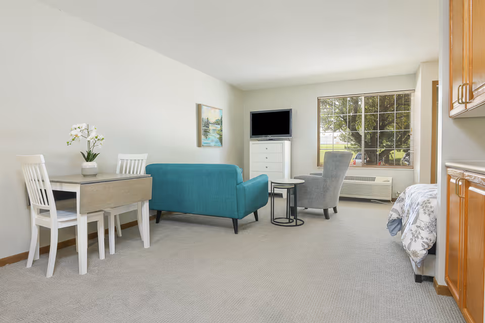 A bright and clean assisted living room featuring a small dining table with two white chairs and a vase with white flowers, a teal loveseat, a gray armchair, a white chest of drawers with a TV on top, a large window with blinds showing a tree outside, and a bed partially visible on the right side next to wooden cabinets.