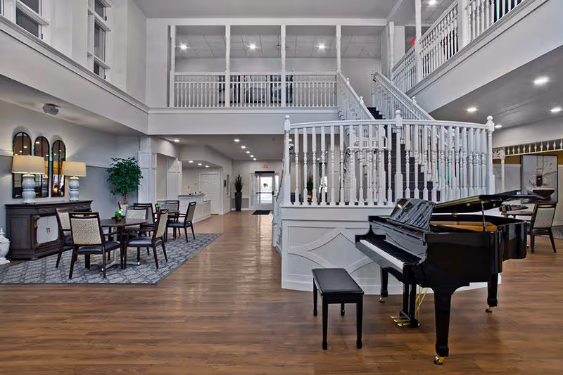 Spacious two-story interior common area with a grand piano, seating tables, and a white staircase and balcony.
