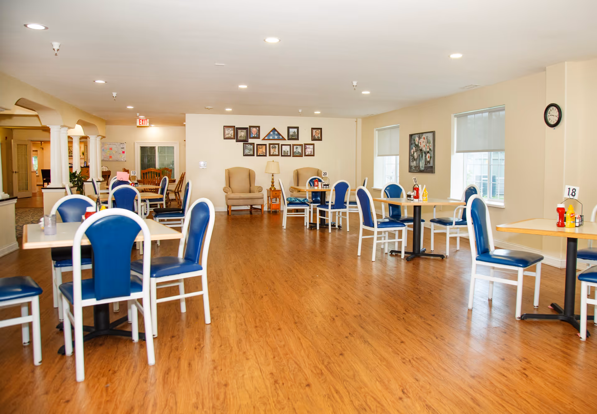 A bright dining room with several tables and blue cushioned chairs arranged neatly on a wooden floor. The room has cream-colored walls with framed pictures and a clock, and two beige armchairs are placed against the far wall. Windows with blinds allow natural light to enter the space.