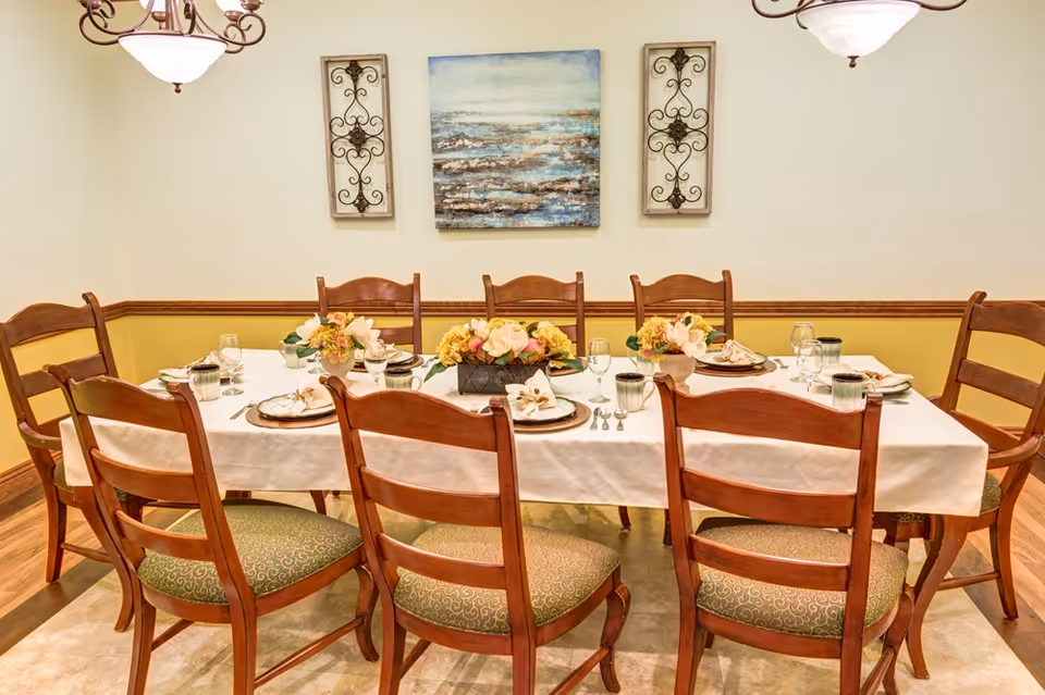 Formal dining room with a long set table, wooden chairs, floral centerpieces, and wall artwork.