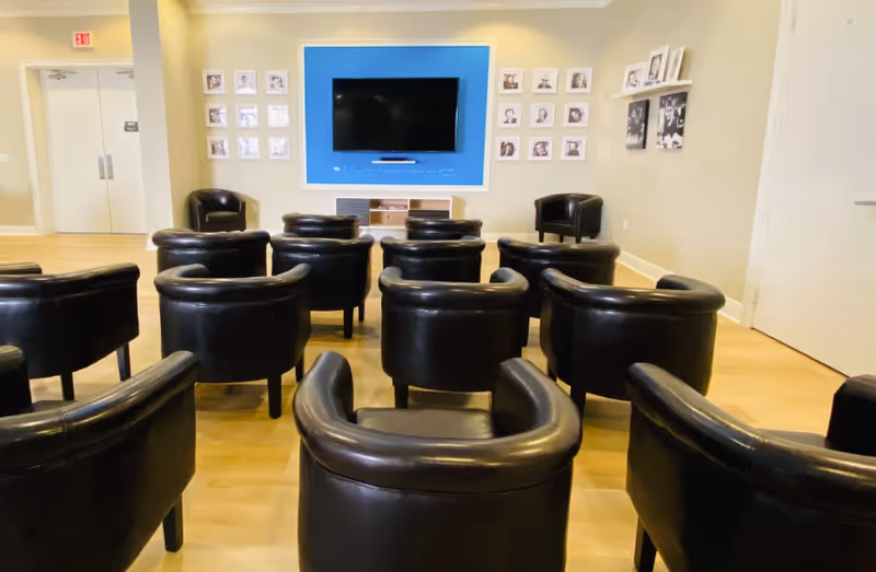 A small auditorium or meeting room with several black leather chairs arranged in rows facing a wall-mounted flat screen TV. The TV is set against a blue accent wall with framed photos on either side. The room has light-colored wooden flooring and neutral-colored walls.