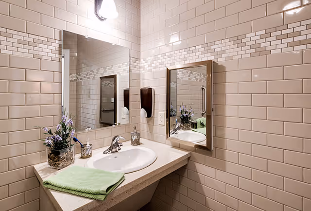 A tiled bathroom vanity with a sink, mirror, soap dispenser, decorative flowers, and a folded green towel.