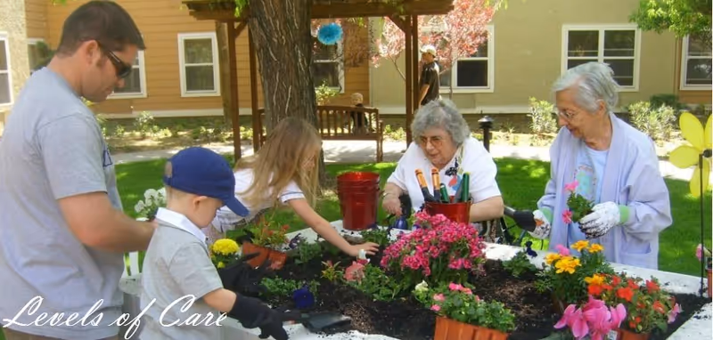 Two elderly women and two children gardening together outdoors at a raised flower bed with various colorful flowers and gardening tools, with a man standing nearby and a building in the background.