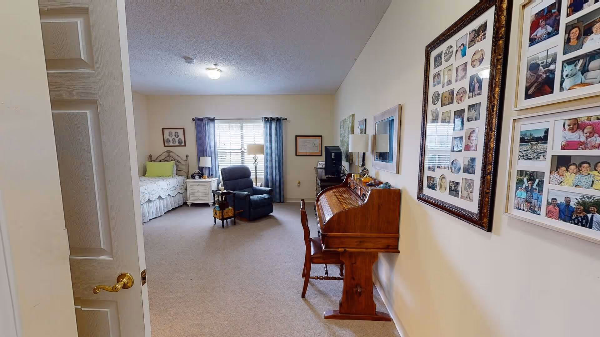 Interior view of a senior living facility room with a bed, a recliner chair, a side table with a lamp, a wooden roll-top desk with a chair, and framed photo collages on the wall. The room has beige walls and carpet, with a window covered by blue curtains letting in natural light.
