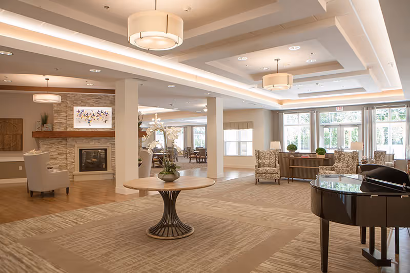 Spacious and well-lit common area in a senior living facility featuring a round table with a flower arrangement in the center, comfortable armchairs, a fireplace with artwork above it, large windows letting in natural light, and a black grand piano on the right side.