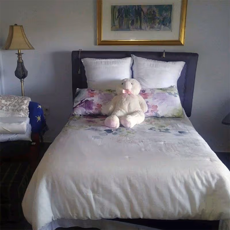 A neatly made bed with white and floral bedding, two white pillows, and a stuffed teddy bear with a pink bow sitting in the center. To the left of the bed is a lamp on a side table and a folded blanket on a chair. A framed painting hangs on the wall above the bed.