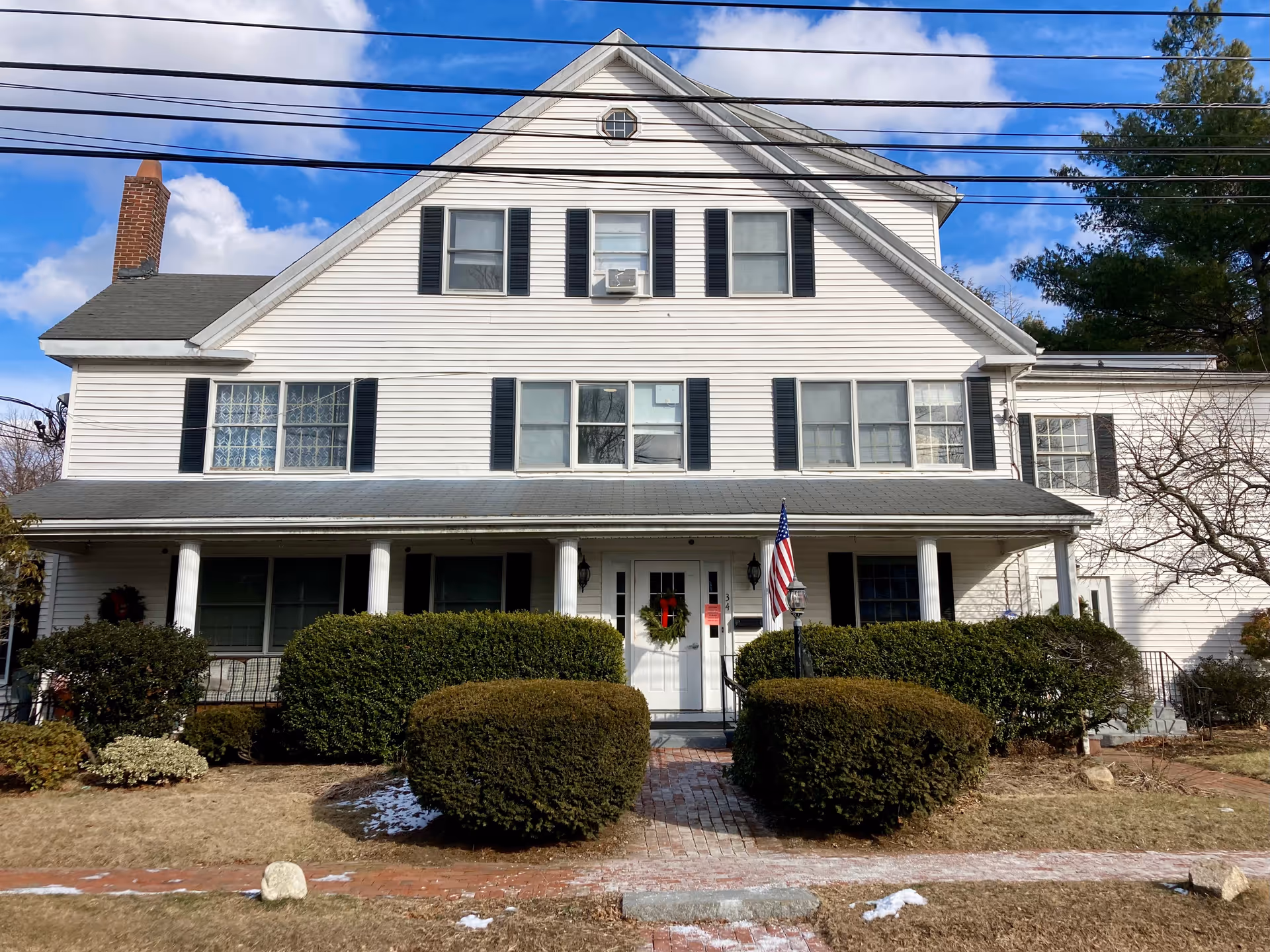 Front facade of a white, multi-story colonial-style house with a covered porch, trimmed shrubs, and an American flag.