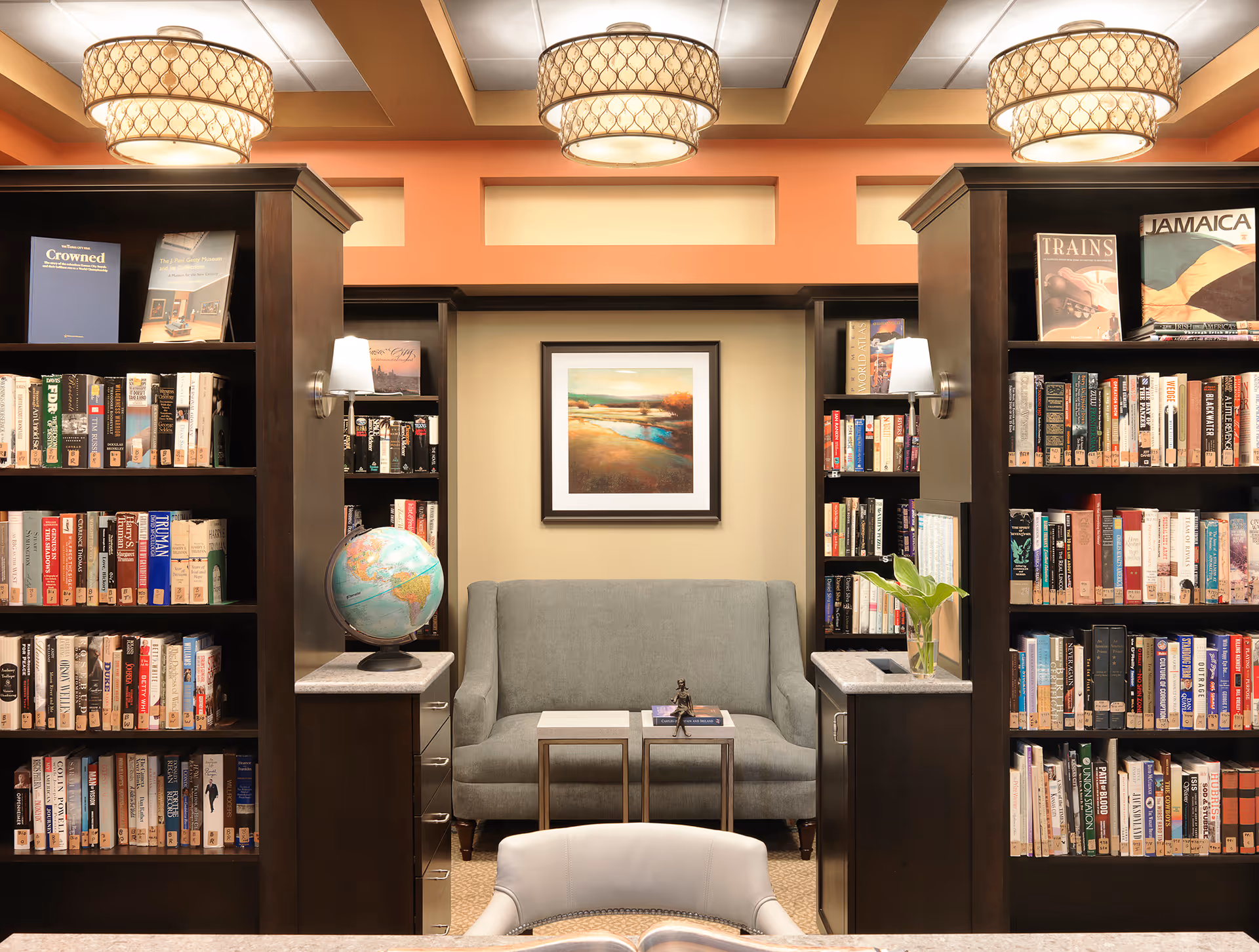 A cozy library room with dark wooden bookshelves filled with books on either side. In the center, there is a gray upholstered loveseat with two small tables in front of it, one holding a book and a small statue. A globe sits on a countertop to the left, and a small plant in a glass vase is on the right countertop. The walls are painted in warm tones with a framed landscape painting above the loveseat. Three decorative ceiling lights illuminate the room.