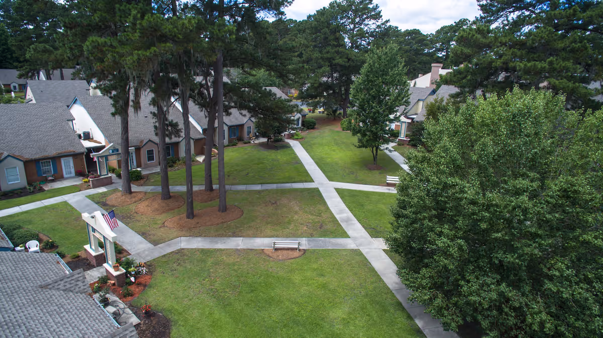 Aerial view of a grassy courtyard with intersecting concrete walkways, benches, tall trees, and surrounding single-story senior living cottages.