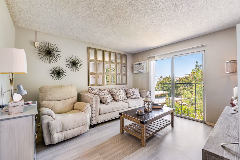 Bright living room with a beige sofa and recliner, wooden coffee table, decorative wall art, and a sliding glass door to a balcony.