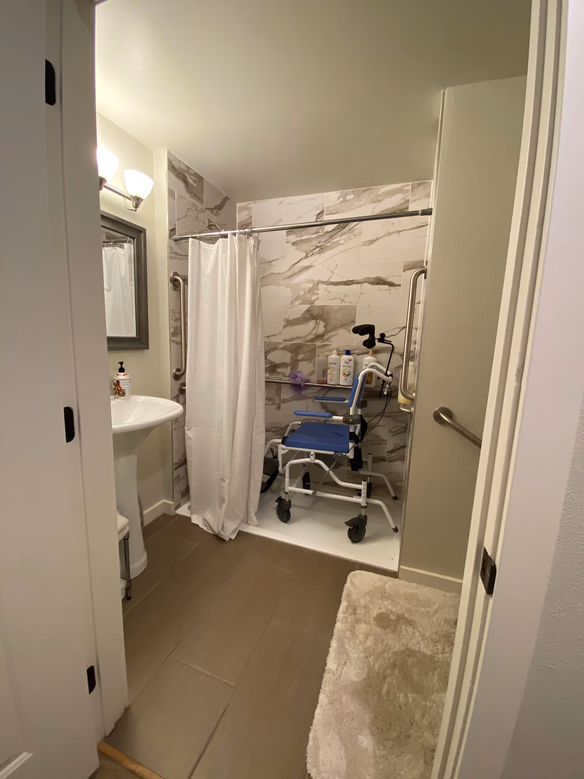 A small bathroom with a white pedestal sink, a mirror above it, and a shower area with marble-patterned tiles. The shower has a white curtain and contains a blue shower chair with wheels and safety grab bars on the walls. There are various bottles of toiletries on a shelf in the shower. A beige bath mat is on the floor outside the shower.