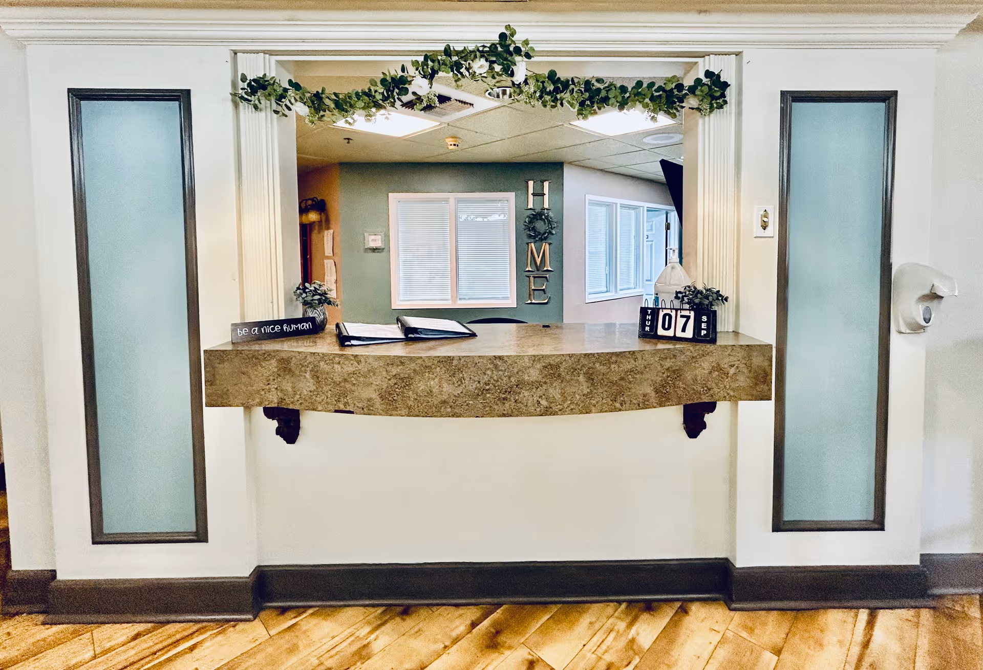 Reception desk in a senior living facility with a decorative counter, greenery, and a 'HOME' sign on the back wall.