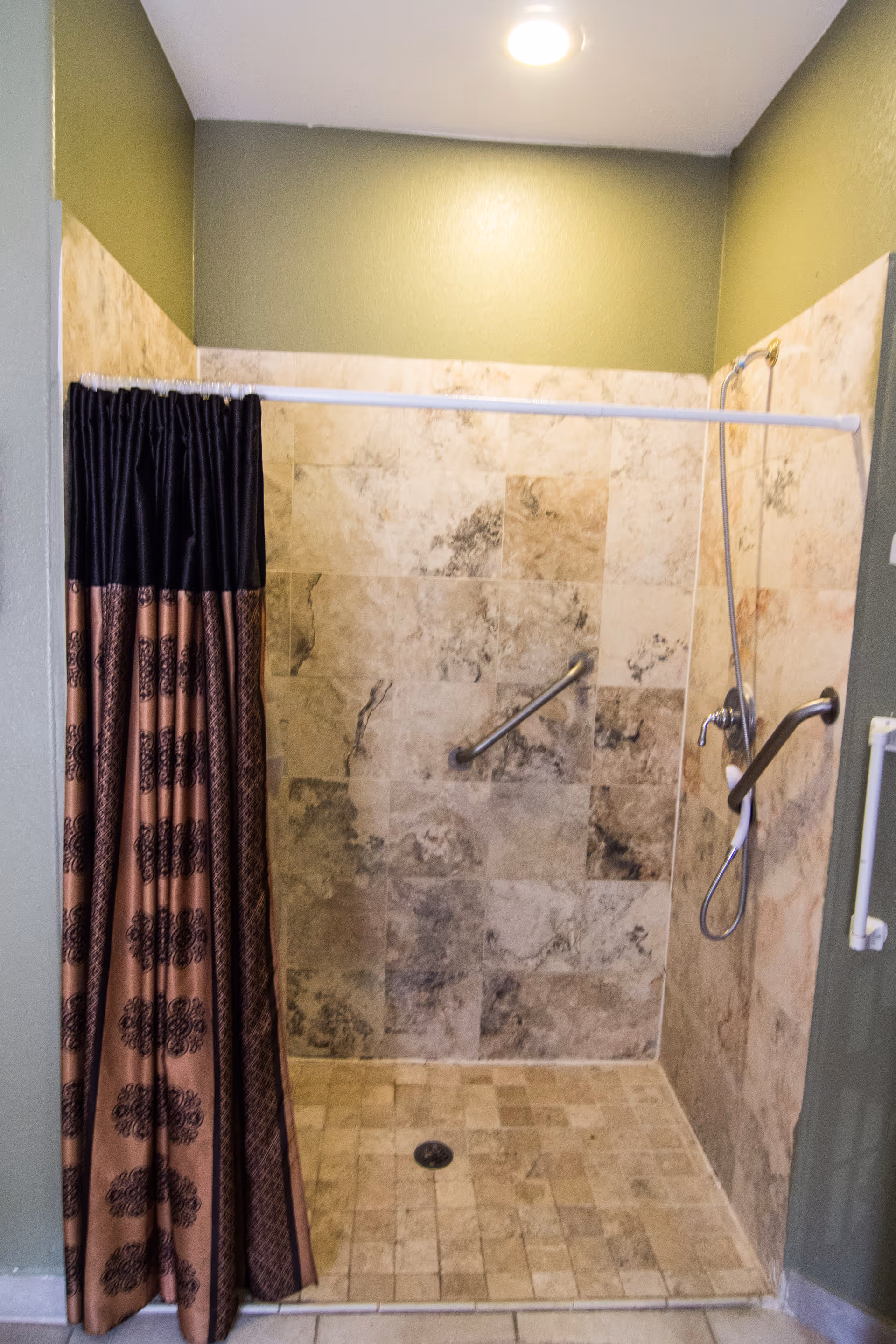 A tiled walk-in shower with beige and brown marbled tiles on the walls and floor. There is a shower curtain with a black and brown pattern partially drawn to the left. The shower has two metal grab bars for support and a handheld showerhead attached to the wall. The walls outside the shower are painted green, and a ceiling light illuminates the area.