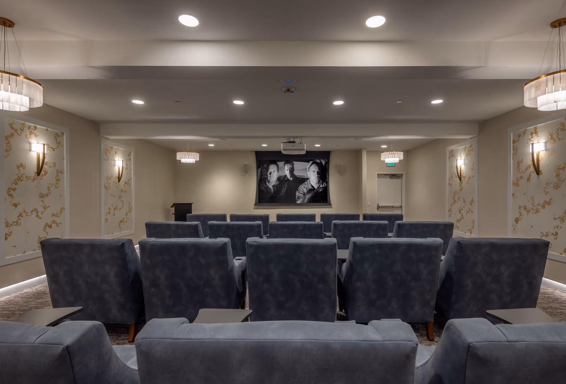 A small theater room with multiple rows of comfortable dark gray armchairs facing a large screen displaying a black and white image of three men. The room has soft lighting with wall sconces and ceiling lights, decorative wall panels with gold patterns, and a podium near the front.