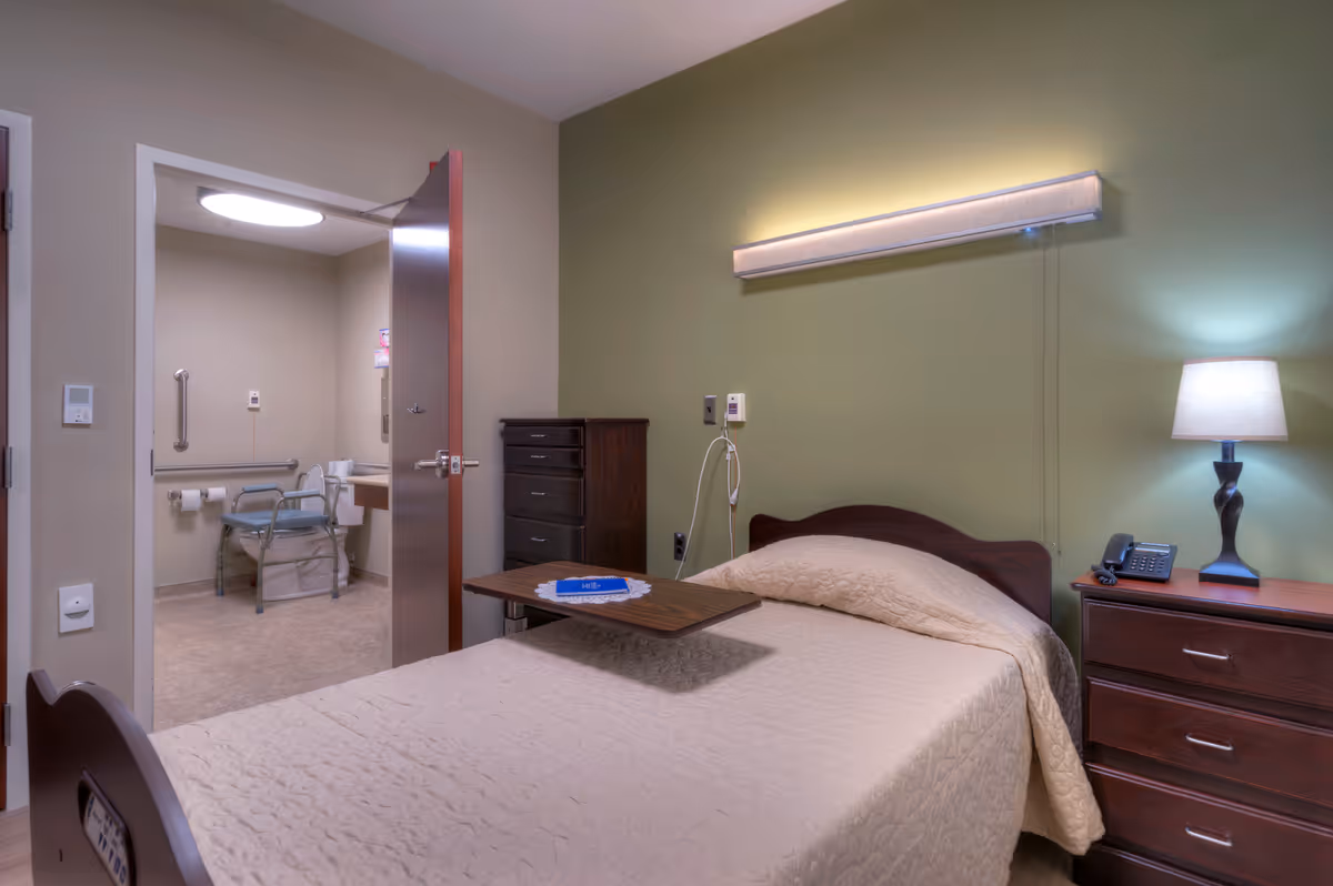 A furnished single bedroom in a senior facility with a bed, bedside table and lamp, dresser, and an open door showing an accessible bathroom.