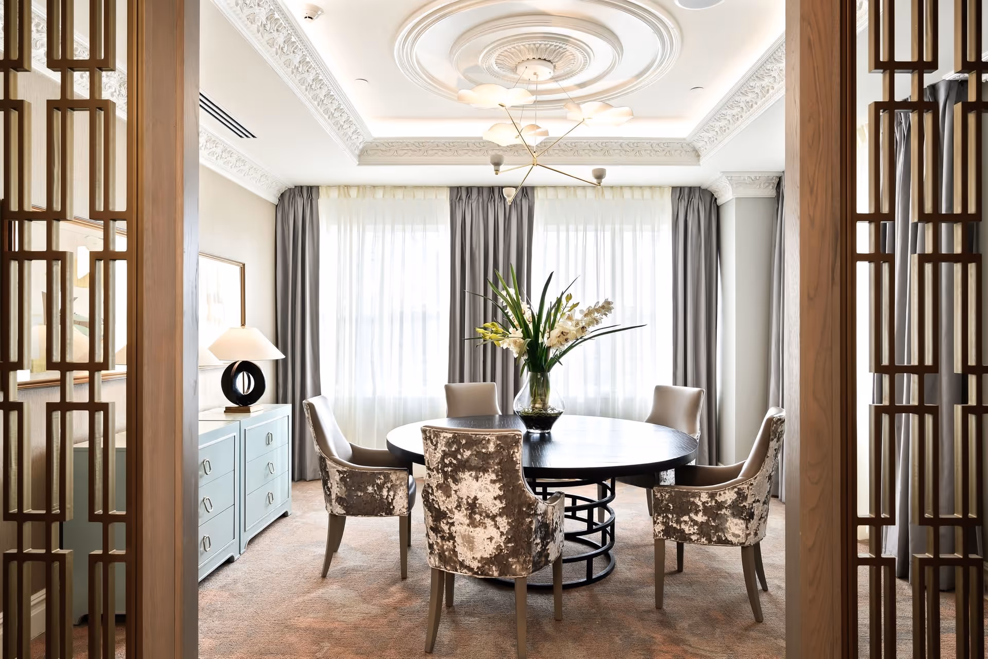 A bright dining room with a round black table surrounded by six upholstered chairs, some with a patterned fabric and others in solid beige. A vase with white flowers sits in the center of the table. The room features large windows with sheer white curtains and gray drapes, ornate crown molding on the ceiling, a modern chandelier, and a light blue sideboard with a lamp on top. Wooden decorative panels frame the entrance to the room.