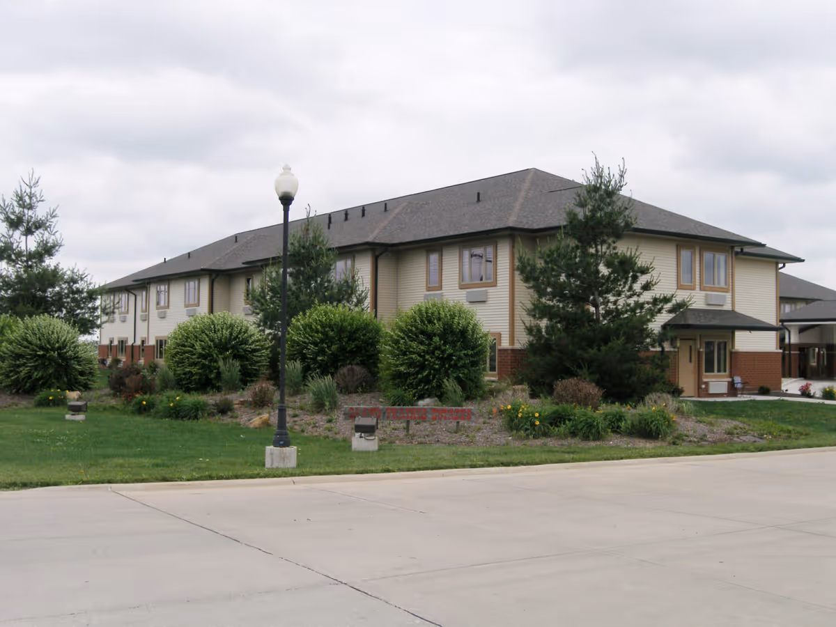 Two-story senior living building with landscaped shrubs, a lamppost, and a cloudy sky.