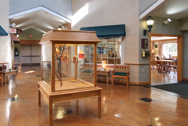 Interior view of a senior living facility lobby area with a wooden display case containing birdhouses in the center. There are wooden benches with green cushions along the walls, a table with a lamp, and a dining area visible through an open doorway on the right. The floor is polished and the ceiling has recessed lighting.