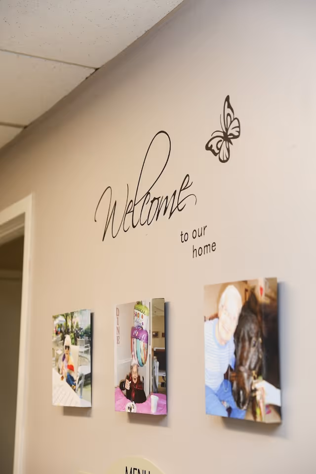 A beige wall with the words 'Welcome to our home' written in decorative black script along with a butterfly graphic. Below the text are three framed photos showing elderly people engaged in various activities, including sitting outdoors, celebrating a birthday, and interacting with a horse.